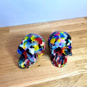 May include: Two colourful painted ceramic skull figurines. The skulls are painted in a variety of bright colours, including red, yellow, blue, green, and purple. The skulls are both facing forward, and the paint is splattered and textured.