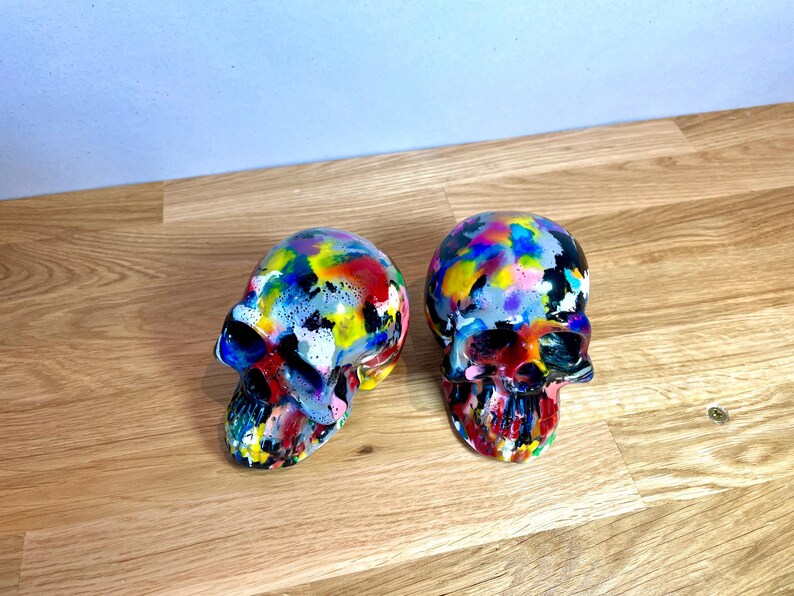 May include: Two colourful painted ceramic skull figurines. The skulls are painted in a variety of bright colours, including red, yellow, blue, green, and purple. The skulls are both facing forward, and the paint is splattered and textured.