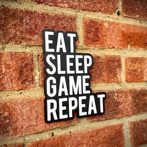 May include: A black and white sign with the words "EAT SLEEP GAME REPEAT" in a stacked format. The sign is mounted on a red brick wall, with the brick texture visible in the background.