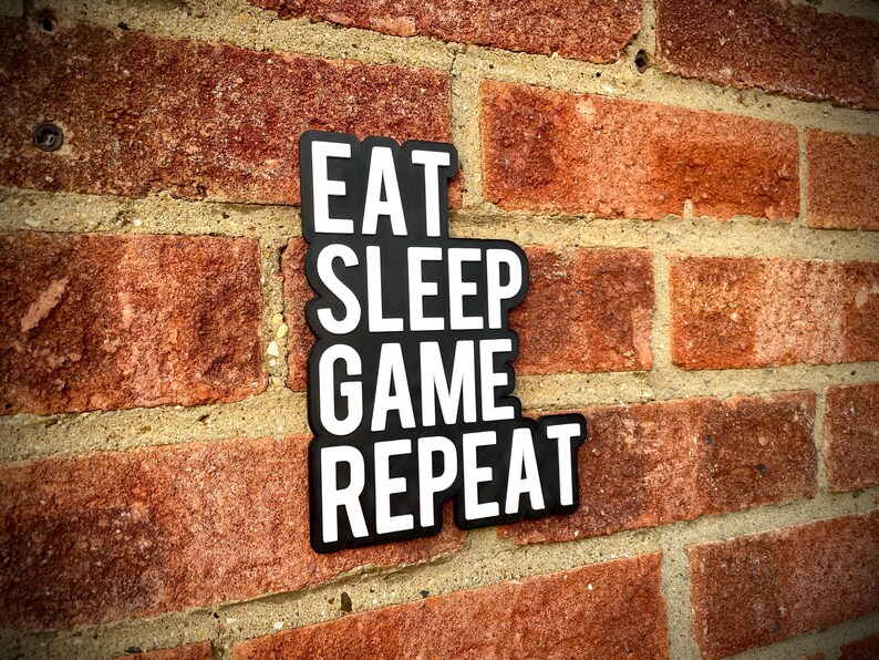 May include: A black and white sign with the words "EAT SLEEP GAME REPEAT" in a stacked format. The sign is mounted on a red brick wall, with the brick texture visible in the background.