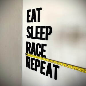 May include: Black letters spell "EAT SLEEP RACE REPEAT" on a white wall. A yellow tape measure is placed horizontally across the words. The tape measure shows measurements in inches.