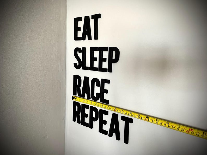 May include: Black letters spell "EAT SLEEP RACE REPEAT" on a white wall. A yellow tape measure is placed horizontally across the words. The tape measure shows measurements in inches.