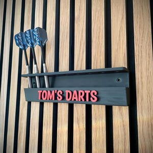 May include: A dark gray dart holder with the text "TOM'S DARTS" in red, mounted on a wooden wall. Three darts with black shafts and blue and black flights are in the holder. The wall has vertical wood panels.