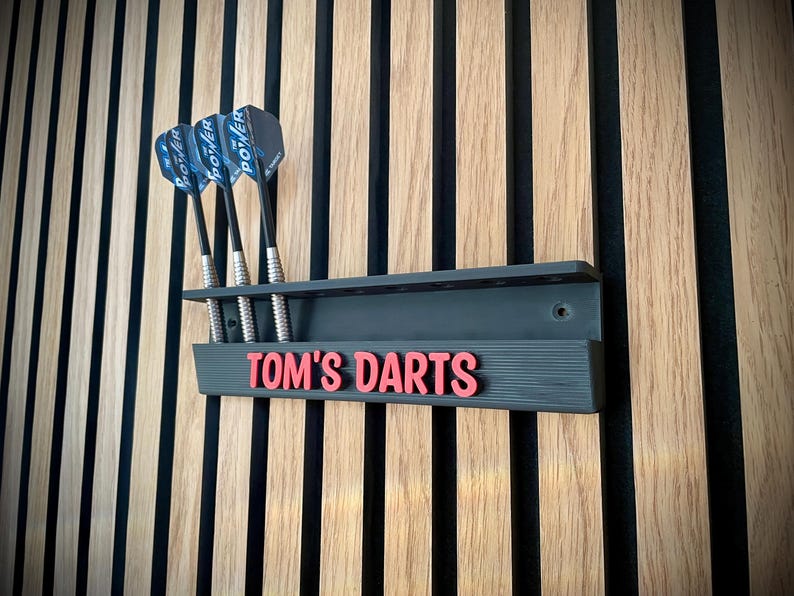 May include: A dark gray dart holder with the text "TOM'S DARTS" in red, mounted on a wooden wall. Three darts with black shafts and blue and black flights are in the holder. The wall has vertical wood panels.