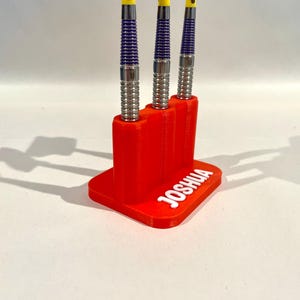 Op de afbeelding: Een rode 3D-geprinte darthouder met de naam "JOSHUA" in het wit. Drie darts met zilveren, paarse en gele accenten worden weergegeven. De darts staan verticaal en worden door de houder vastgehouden.
