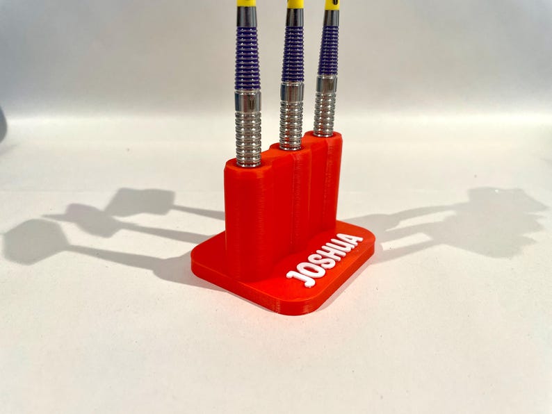 Op de afbeelding: Een rode 3D-geprinte darthouder met de naam "JOSHUA" in het wit. Drie darts met zilveren, paarse en gele accenten worden weergegeven. De darts staan verticaal en worden door de houder vastgehouden.