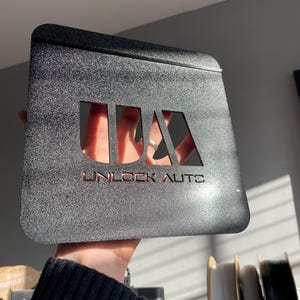 May include: A black, textured metal plate with the text "UNLOCK AUTOS" cut out. The plate has a rectangular shape with rounded corners and is held up in front of a light background.