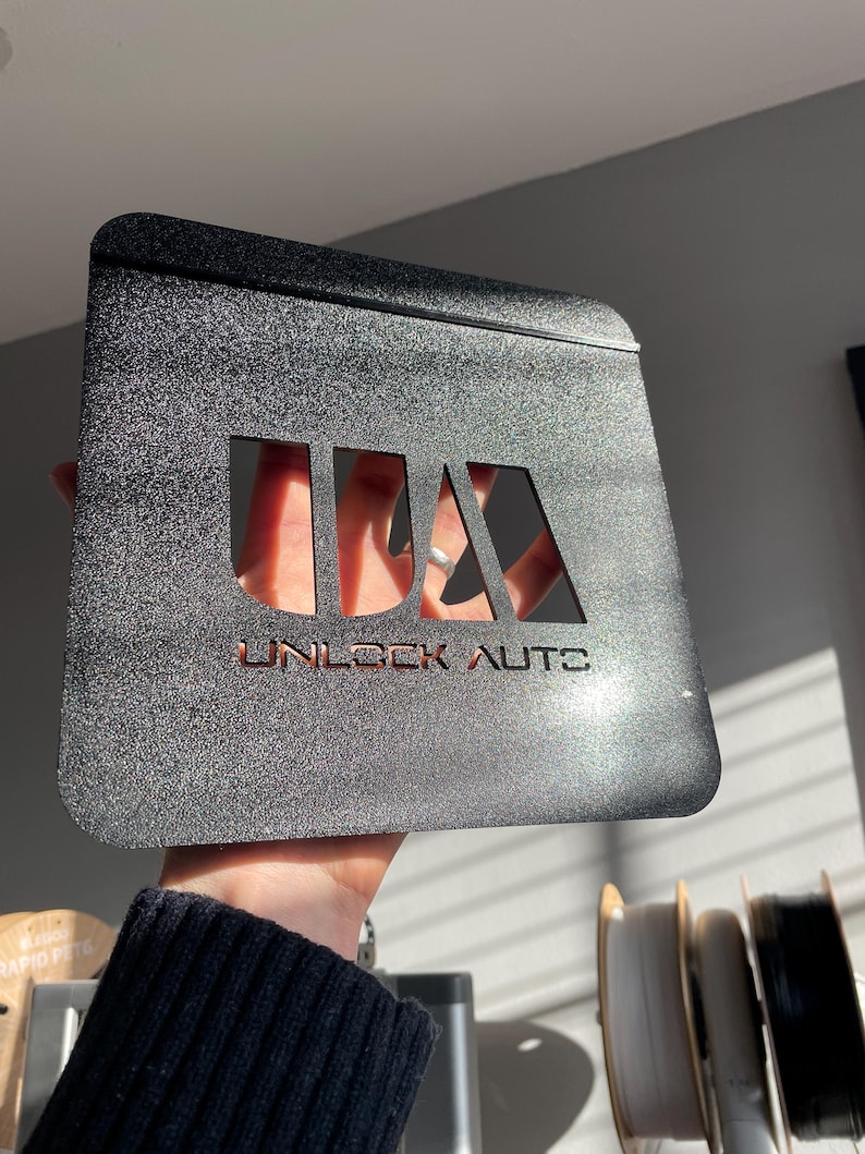 May include: A black, textured metal plate with the text "UNLOCK AUTOS" cut out. The plate has a rectangular shape with rounded corners and is held up in front of a light background.