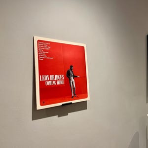 May include: A red vinyl record album cover with a black and white photo of a man in a suit and tie. The album is titled "Leon Bridges Coming Home" and lists the songs "Coming Home", "Better Man", "Brown Skin Girl", "Smooth Sailin", "Shine", "Lisa Sawyer", "Flowers", "Pull Away", "Twistin' & Groovin", "River".