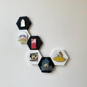 May include: A collection of Disney-themed pins displayed on a white wall. The pins are attached to black and white hexagon-shaped holders. The pins feature characters and logos from Disney films and parks, including Disneyland and The Walt Disney Family Museum.