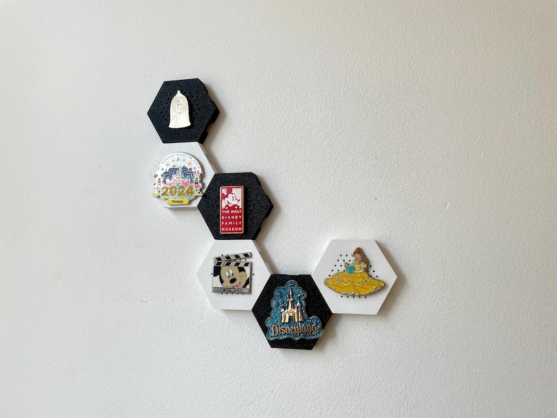 May include: A collection of Disney-themed pins displayed on a white wall. The pins are attached to black and white hexagon-shaped holders. The pins feature characters and logos from Disney films and parks, including Disneyland and The Walt Disney Family Museum.