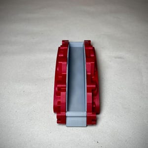 May include: A red and gray 3D-printed object, possibly a component or part. The object has a rectangular shape with a central gray channel and red, textured sides. The design appears to be symmetrical.