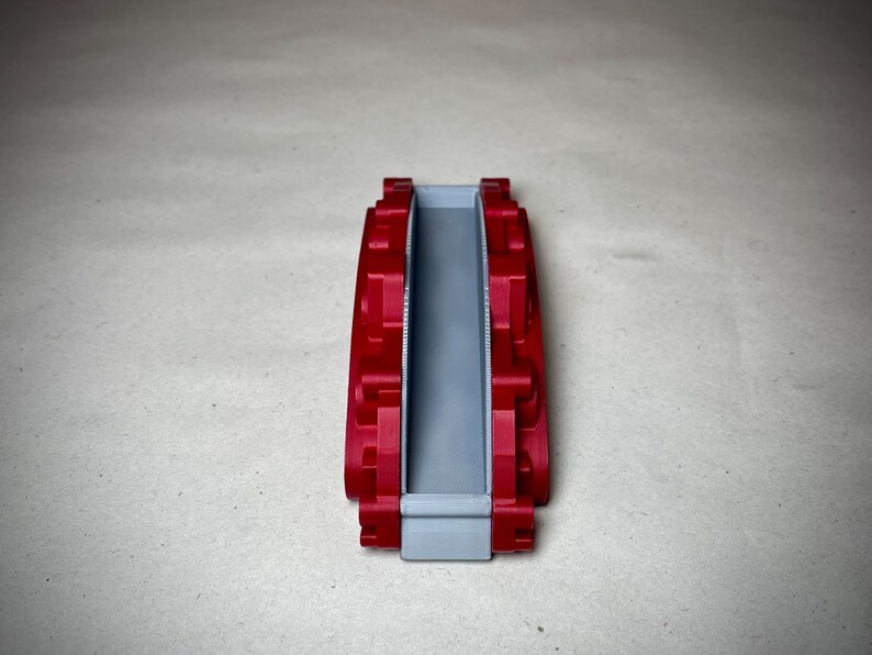 May include: A red and gray 3D-printed object, possibly a component or part. The object has a rectangular shape with a central gray channel and red, textured sides. The design appears to be symmetrical.