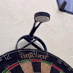 Dart board with attached lamp on the top. The lamp extends out in front of the board in order to cast light on the play area. Lamp is black modern design with a puck light at the end.