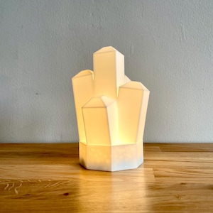 Pu&ograve; includere: Una lampada in ceramica bianca a forma di grappolo di cristalli. La lampada &egrave; accesa ed emana una luce calda.