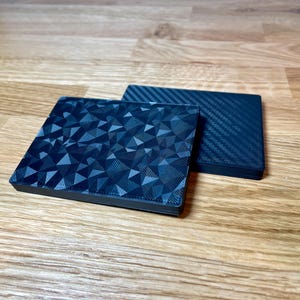 May include: Two black square coasters. One coaster has a geometric pattern and the other has a carbon fiber pattern.