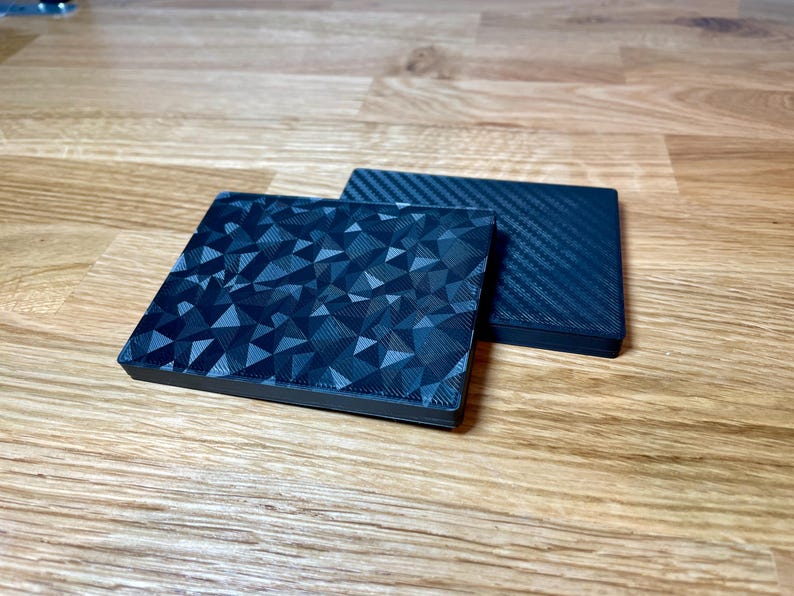 May include: Two black square coasters. One coaster has a geometric pattern and the other has a carbon fiber pattern.