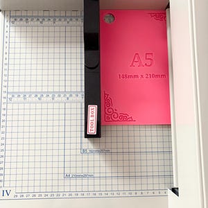 May include: A white paper cutter with a black handle and a grid-marked base. A bright pink A5 sheet measuring 148mm x 210mm is positioned under the black cutting arm. The base features measurement markings and the text HFS HardwareFactoryStore.com.