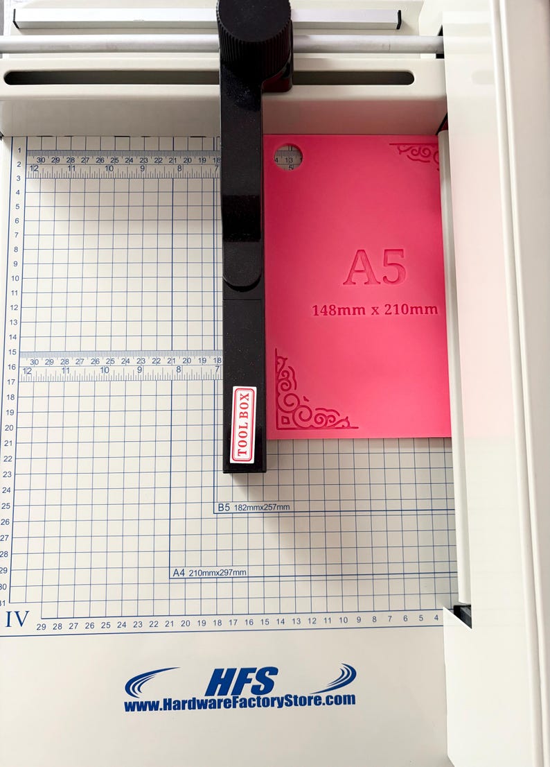May include: A white paper cutter with a black handle and a grid-marked base. A bright pink A5 sheet measuring 148mm x 210mm is positioned under the black cutting arm. The base features measurement markings and the text HFS HardwareFactoryStore.com.