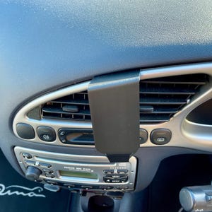 May include: A black phone mount attached to the air vent of a car's dashboard. The mount is designed to hold a smartphone securely in place. The car's radio and climate control buttons are visible in the background.