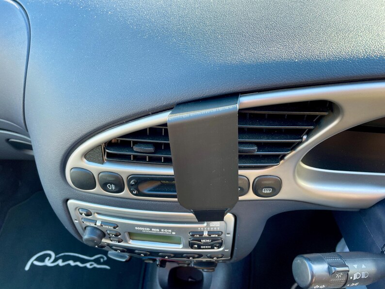 May include: A black phone mount attached to the air vent of a car's dashboard. The mount is designed to hold a smartphone securely in place. The car's radio and climate control buttons are visible in the background.