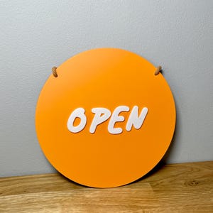 May include: A round orange sign with the word "OPEN" in white letters. The sign has a rope attached to it for hanging.