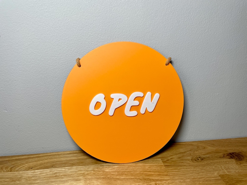 May include: A round orange sign with the word "OPEN" in white letters. The sign has a rope attached to it for hanging.