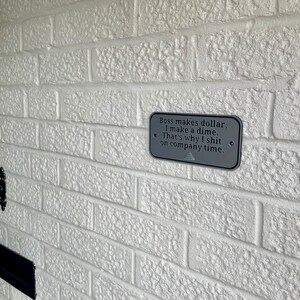 May include: A rectangular grey metal sign with the text "Boss makes dollar, I make a dime. That's why I shit on company time." is mounted on a white brick wall. The sign has two small holes on each side.