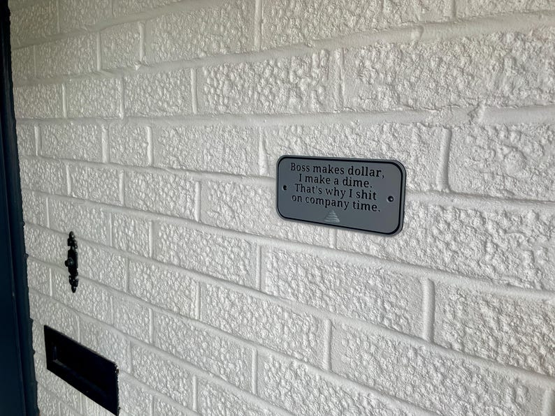 May include: A rectangular grey metal sign with the text "Boss makes dollar, I make a dime. That's why I shit on company time." is mounted on a white brick wall. The sign has two small holes on each side.