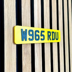 May include: A yellow rectangular sign with the text "W965 RDU" in dark teal lettering. The sign is mounted on a wooden wall with vertical wood panels and black stripes.