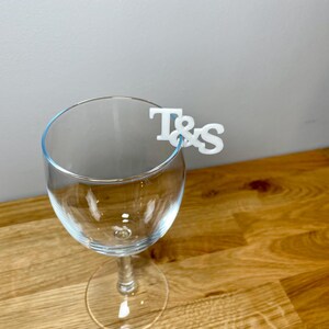 Wine glass with a set of initial letters clipped onto the side. Drink topper is white and the photo shows a grey background.