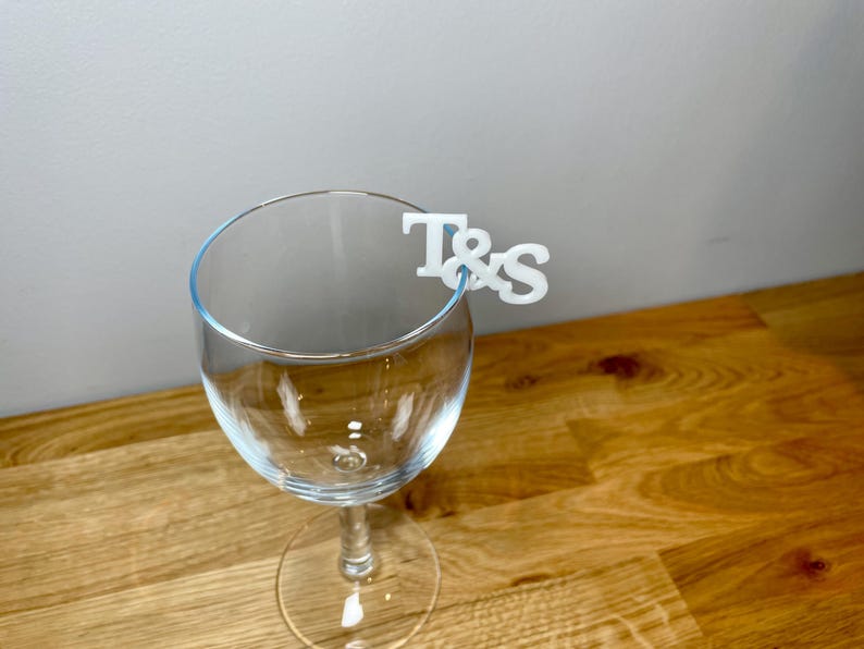 Wine glass with a set of initial letters clipped onto the side. Drink topper is white and the photo shows a grey background.