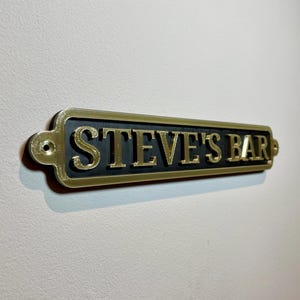 May include: A gold-colored metal sign with black lettering that reads "STEVE'S BAR".