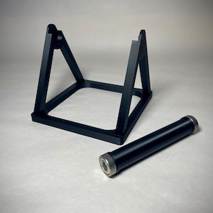 May include: Black metal stand with a square base and two angled supports. A black cylindrical object with silver ends rests beside the stand. The objects are on a light surface.