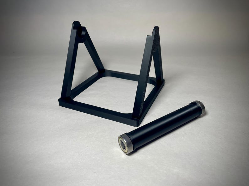 May include: Black metal stand with a square base and two angled supports. A black cylindrical object with silver ends rests beside the stand. The objects are on a light surface.