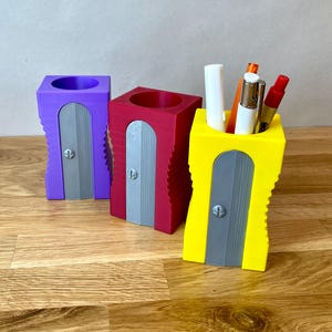 May include: Three pencil sharpeners, one purple, one red, and one yellow, with a pencil holder on top of the yellow sharpener.