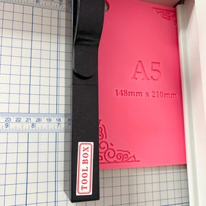 May include: A black paper trimmer with a vertical handle and a white label reading TOOL BOX, positioned over a bright pink A5 plastic sheet measuring 148mm x 210mm, set against a white cutting mat with blue grid lines and imperial and metric rulers.