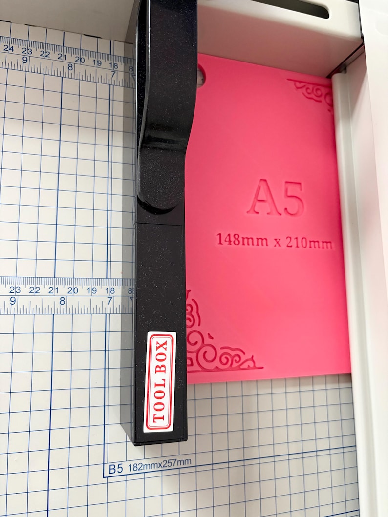May include: A black paper trimmer with a vertical handle and a white label reading TOOL BOX, positioned over a bright pink A5 plastic sheet measuring 148mm x 210mm, set against a white cutting mat with blue grid lines and imperial and metric rulers.