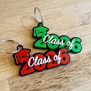 May include: Two keychains with the text "Class of 2026". One keychain is red with a black outline and a graduation cap, the other is green with a black outline and a graduation cap. Both have silver key rings.