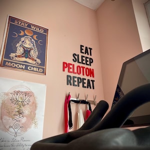 May include: A room with motivational wall art and exercise equipment. A poster reads "Stay Wild Moon Child" and another features a lotus flower. The words "Eat Sleep Peloton Repeat" are on the wall. A stationary bike is in the foreground.