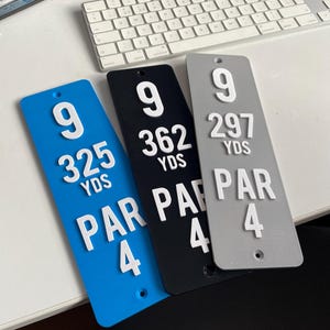 May include: Three golf course hole markers in blue, black, and gray. Each marker displays the number 9, yardage, and Par 4 in white raised lettering. The blue marker reads 325 YDS, the black 362 YDS, and the gray 297 YDS.