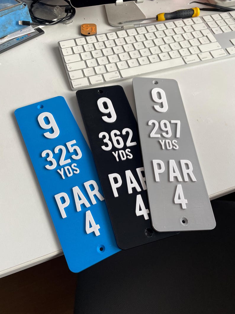 May include: Three golf course hole markers in blue, black, and gray. Each marker displays the number 9, yardage, and Par 4 in white raised lettering. The blue marker reads 325 YDS, the black 362 YDS, and the gray 297 YDS.