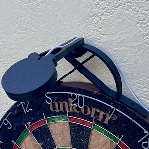 Dart board with attached lamp on the top. The lamp extends out in front of the board in order to cast light on the play area. Lamp is black modern design with a puck light at the end.