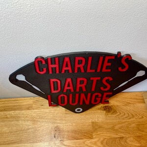 Triangular darts sign with the text charlie's darts lounge in red. Main background is black and has silver inlayed darts logos.