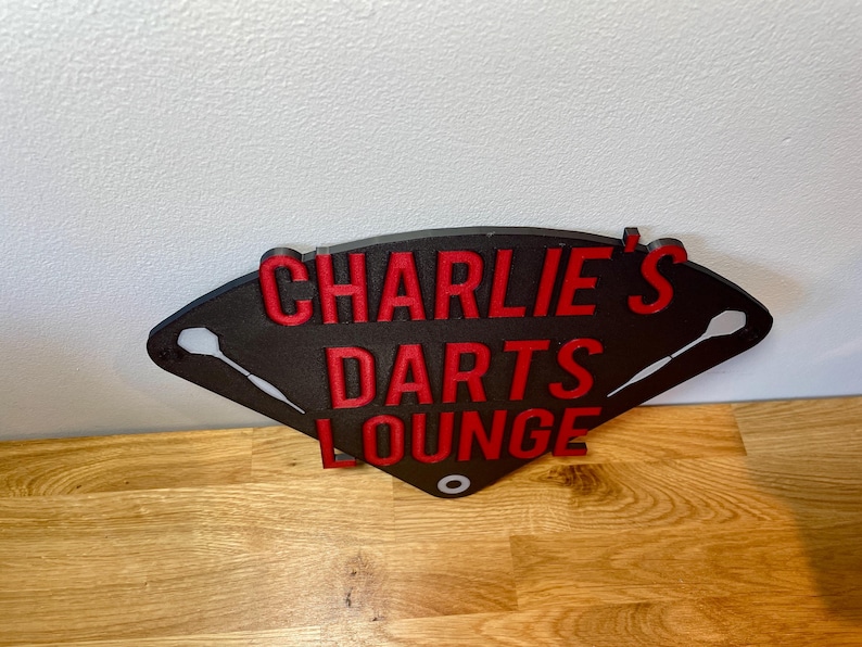 Triangular darts sign with the text charlie's darts lounge in red. Main background is black and has silver inlayed darts logos.