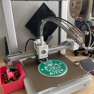 May include: A 3D printer with a green and white Starbucks logo that says "BOO BITCH" printed on the bed. The printer is white and gray, with a pink container holding small, colorful objects. The text "Printthey Spears" is above the printer.
