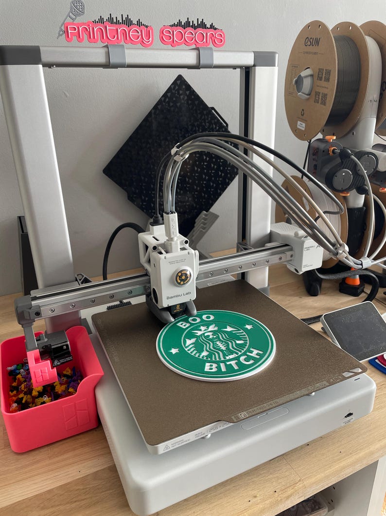 May include: A 3D printer with a green and white Starbucks logo that says "BOO BITCH" printed on the bed. The printer is white and gray, with a pink container holding small, colorful objects. The text "Printthey Spears" is above the printer.