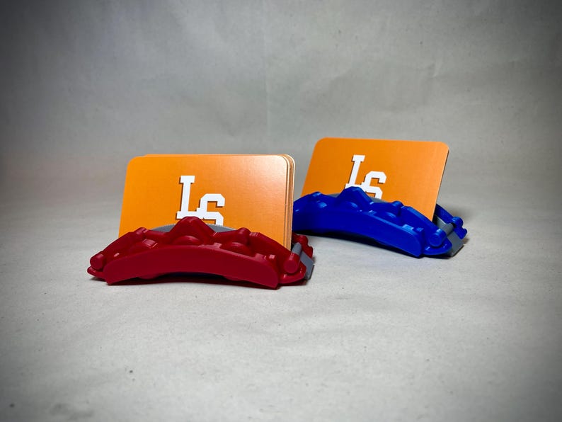 May include: Two card holders, one red and one blue, each supporting an orange card with the white letters "LS". The holders have a textured design. The background is a neutral gray.