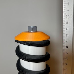 Op de afbeelding: Een wit, zwart en oranje 3D-geprint object met een zeskantige moer bovenop. Het object is gemaakt van meerdere lagen zwart en wit plastic, met een oranje bovenkant. Het object wordt gemeten met een liniaal.
