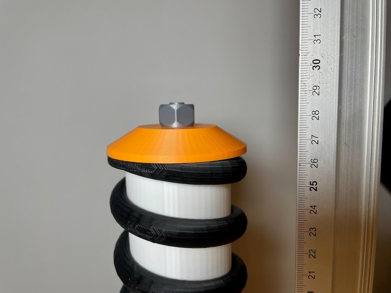 Op de afbeelding: Een wit, zwart en oranje 3D-geprint object met een zeskantige moer bovenop. Het object is gemaakt van meerdere lagen zwart en wit plastic, met een oranje bovenkant. Het object wordt gemeten met een liniaal.